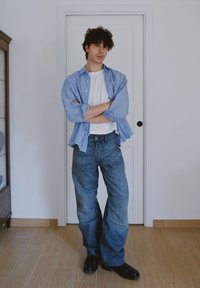 Blå och vit randig skjorta med knappar över en vit t-shirt. Mörkblå överdimensionerade jeans med texturerade paneler och svarta skor. Stående pose.