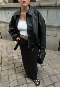 Žena u crnoj kožnoj jakni, bijeloj majici, crnoj suknji, crnim sandalama, koja drži crnu torbu, stoji na kaldrmi.