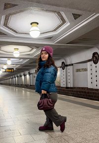 Blågrøn puffjakke, grå bukser og lilla støvler. Bærer en lille lilla håndtaske. Baggrund med en metrostation med dekorativ belysning.