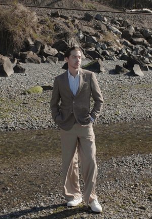 Hombre de pie en la orilla rocosa del río, vestido con una chaqueta marrón, camisa blanca, pantalones beige y zapatos blancos, con las manos en los bolsillos y los ojos cerrados.