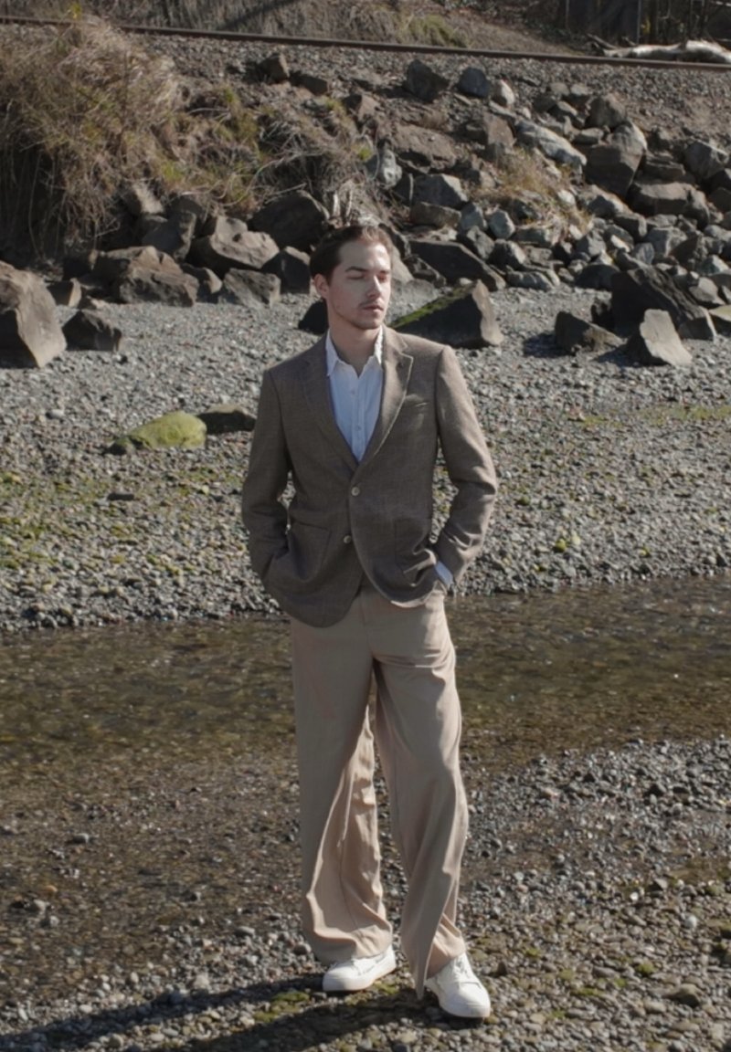 Mann steht am felsigen Flussufer, trägt ein braunes Sakko, weißes Hemd, beige Hose und weiße Schuhe, die Hände in den Taschen, die Augen geschlossen.