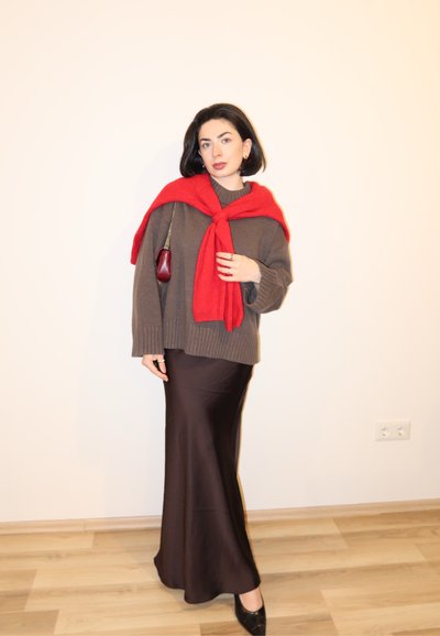 Mujer está de pie en el interior con un suéter marrón, falda larga marrón, suéter rojo sobre los hombros, zapatos negros y lleva un pequeño bolso rojo.