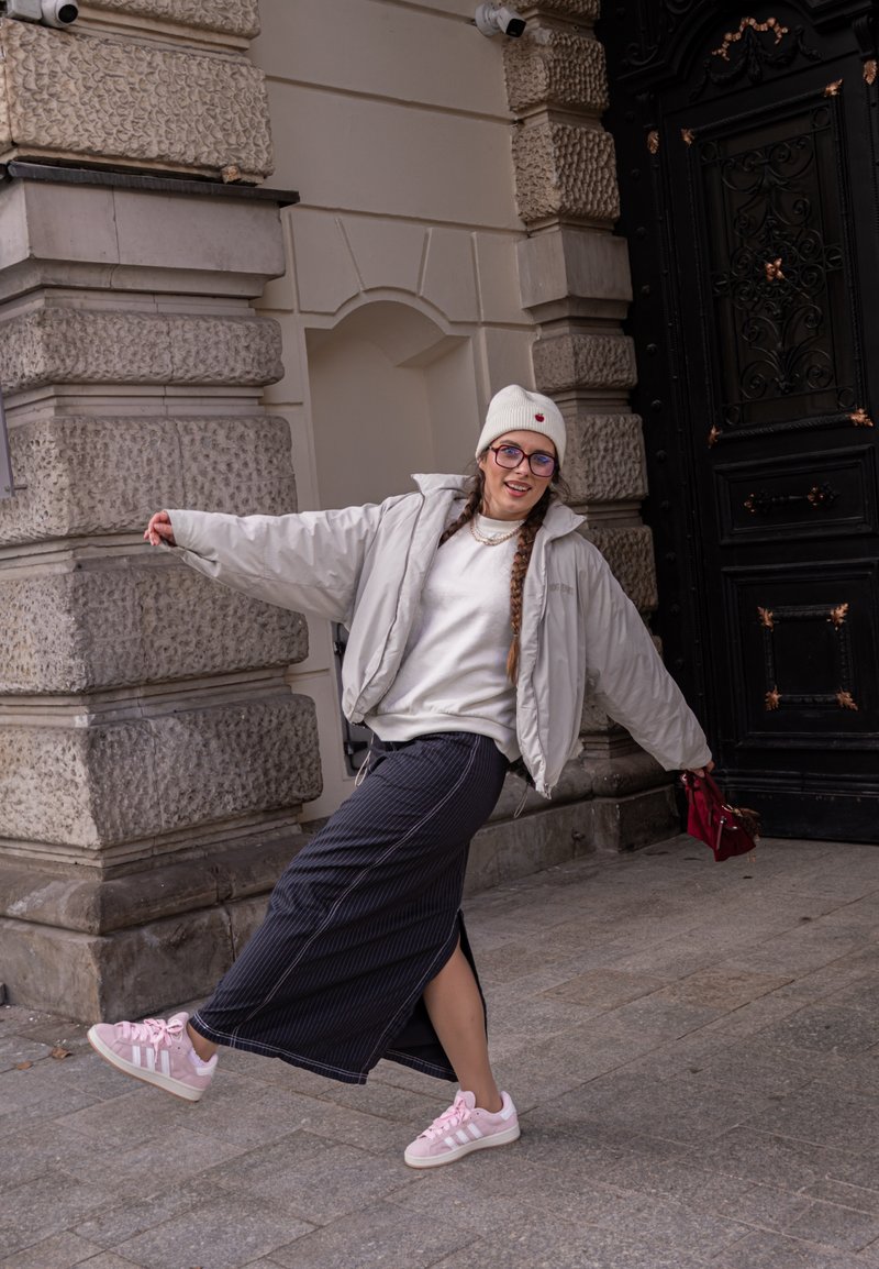 Cilvēks, kurš ģērbies gaiši pelēkā uzpūšamajā jakā, baltā džemperī un garā svītrainā svārku komplektā ar rozā sporta apaviem, stāv pretī teksturētai akmens arhitektūrai.