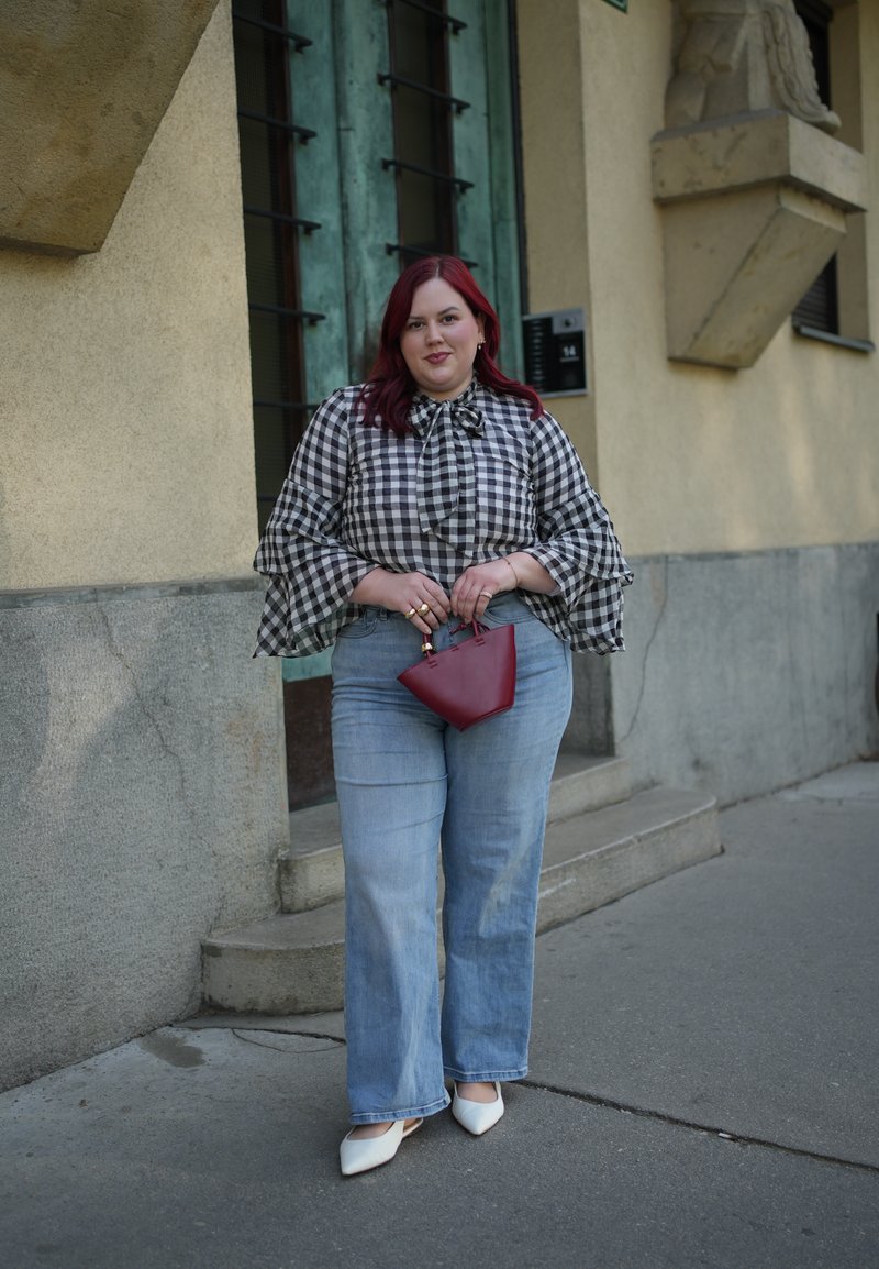 Žena s červenými vlasy nosí černobílou kostkovanou blůzu a světle modré džíny, drží malou červenou kabelku na městském chodníku.