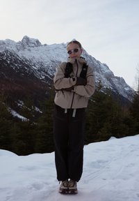 Personne portant des lunettes de soleil et des vêtements d'hiver debout sur la neige devant des montagnes enneigées et des arbres à feuilles persistantes.