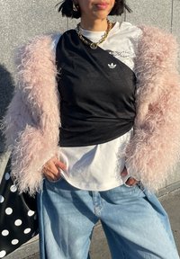 Personne portant une veste rose clair en matière pelucheuse, une chemise blanche avec un superposé en tissu noir, une grosse chaîne en or, un jean bleu ample, et tenant un sac noir à pois.