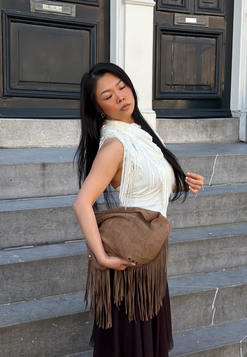 Femme aux longs cheveux noirs portant un haut blanc à franges, tenant un sac marron à franges, debout sur des marches en pierre avec des portes en bois foncé derrière.