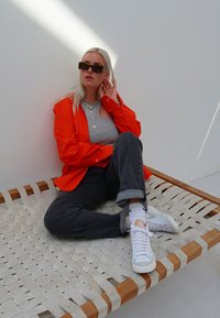 Oranje overhemd met knopen over een grijze T-shirt, donkere jeans, witte sneakers. Textuur bestaat uit een geweven houten zitting en een vlakke muur achtergrond. Zonnebril voegt detail toe.
