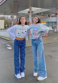 Dwie osoby stoją na stacji benzynowej. Jedna ma na sobie skróconą białą koszulkę z grafiką i niebieskie spodni w kratkę; druga ubrana jest w szary sweter i wyblakłe dżinsy.