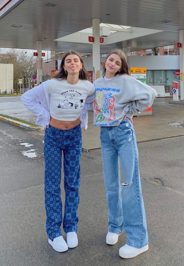 Dos personas se encuentran en una gasolinera. Una lleva una camiseta gráfica blanca recortada y pantalones de cuadros azules; la otra lleva un suéter gris y jeans desgastados.