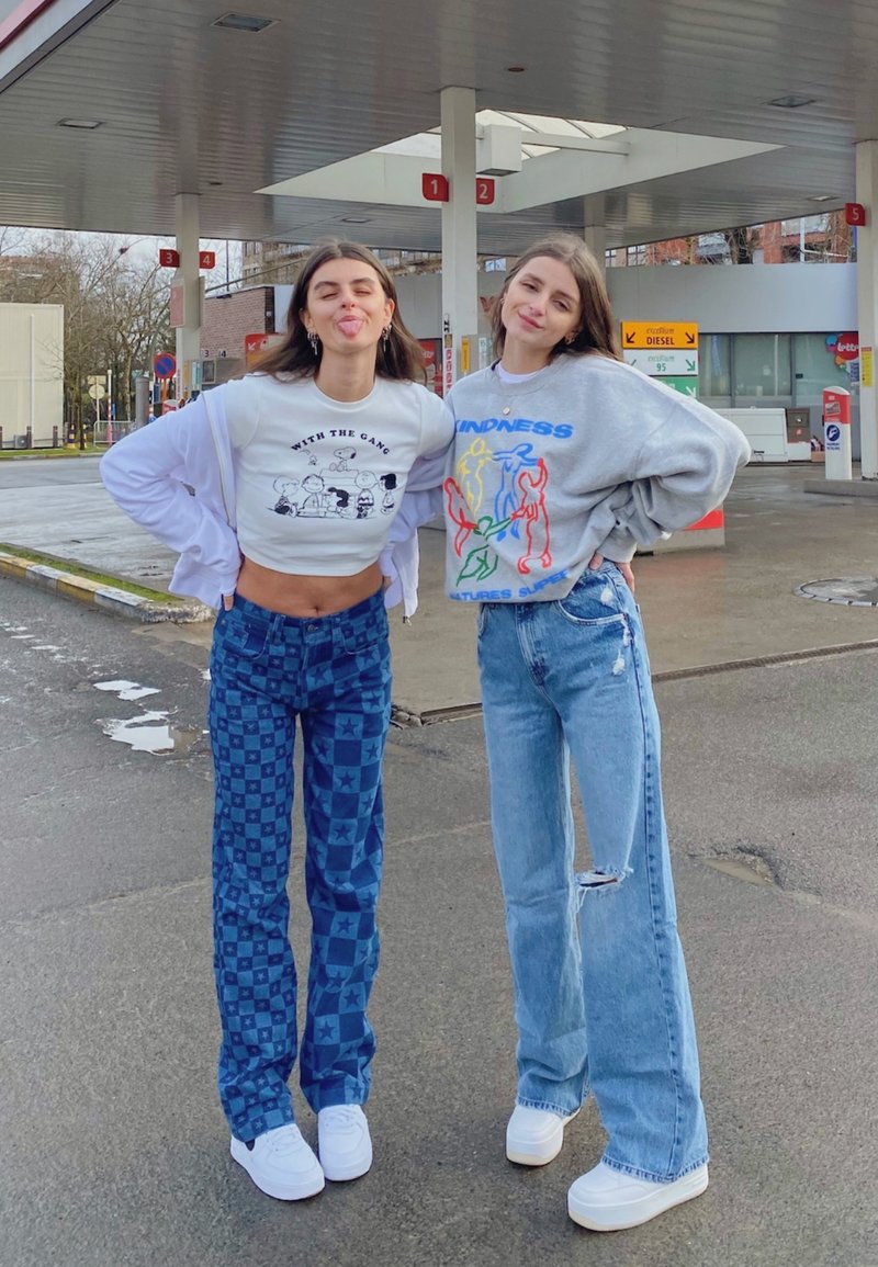 To personer står ved en tankstation. Den ene har en kort hvid grafisk t-shirt og blå ternede bukser på; den anden har en grå sweater og slidte jeans.