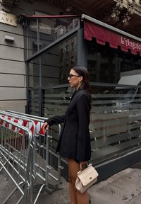 Črn oversized blazer z dolgimi rokavi, v kombinaciji s kratkimi hlačami. Svetlo bež torbica, temna sončna očala in urbana kavarna v ozadju.