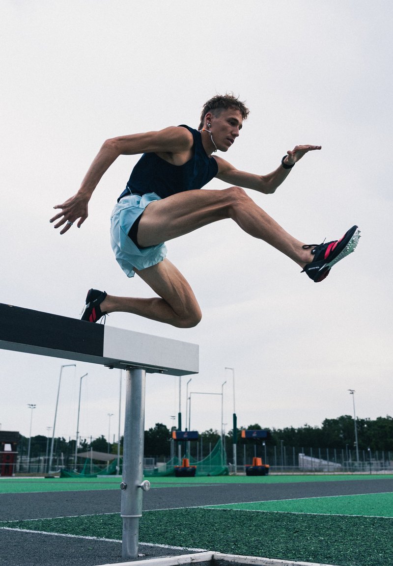 Sportininkas, dėvintis juodą marškinėlį ir šviesiai mėlynas šortus, dienos metu peršoka kliūtį lauke esančioje trasoje.