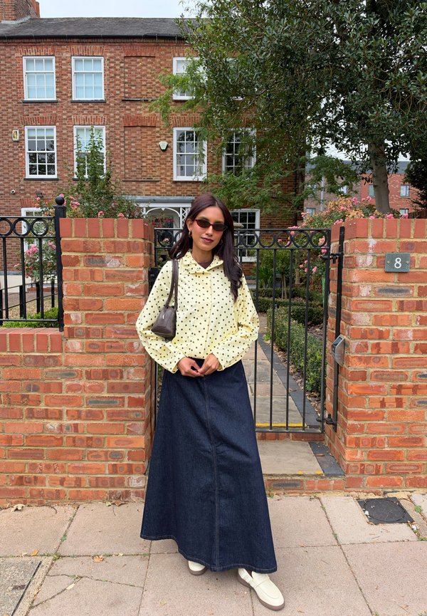 Une femme portant des lunettes de soleil, un chemisier jaune à pois et une longue jupe en jean se tient devant un portail de jardin en briques.