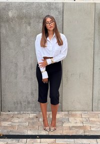 Camisa blanca de botones recortada, pantalones capri negros ajustados y tacones altos con estampado. Cabello largo y liso y gafas visibles. Fondo de hormigón.