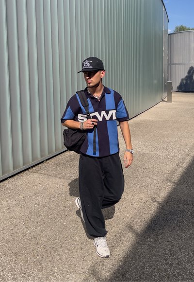 Polo a rayas negras y azules con un gran logotipo gráfico, combinado con pantalones negros sueltos y zapatillas blancas. Gorra negra y mochila.