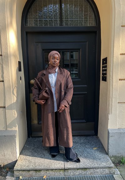 Trench coat marrón, largo, con botones de doble botonadura, vestido sobre una camisa blanca. Pantalones negros y un bolso marrón a juego. Pañuelo en la cabeza de color sólido.