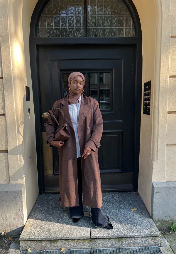Bruine, lange trenchcoat met dubbelzijdige knopen gedragen over een wit shirt. Zwarte broek en een bijpassende bruine handtas. Effen hoofddoek.