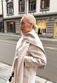 Svetlo bež volneni plašč z velikimi ovratniki in pasom. Mehek material, oversize oblikovanje, v kombinaciji s črno torbico. V ozadju je vidna urbana scenarija.