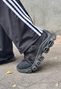 Melnas sporta kurpes ar acs paneļiem, noapaļotu zoli un izteiksmīgu gaisa ventilācijas dizainu. Valkātas kopā ar melnām biksēm ar baltām svītrām.