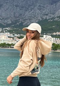 Beige mikina s barevným grafickým potiskem na zádech, doplněná bílou baseballovou čepicí. Dlouhé vlasy spadají přes ramena. Na sobě má tmavé legíny.