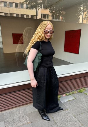 Pantalones anchos negros y una camiseta ajustada negra, complementados con un bolso verde. Llevados con botas negras de punta, con un fondo de galería.