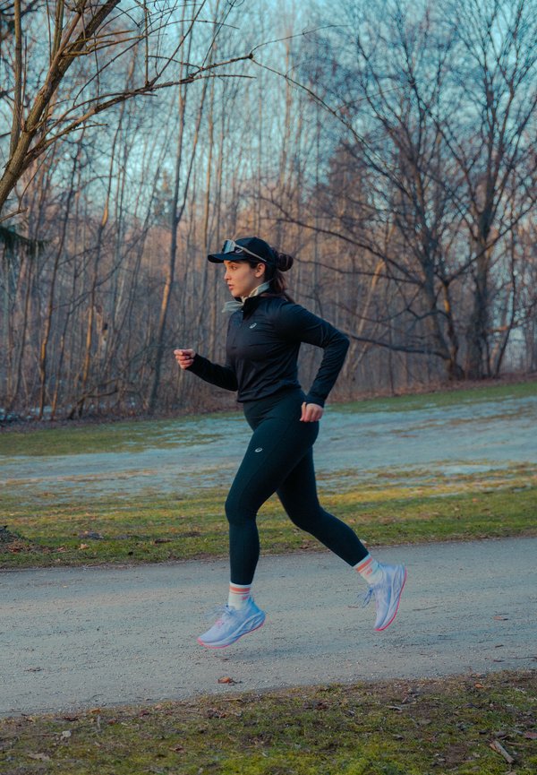Woman in black sportswear and cap jogging along a path through a leafless forest in the early morning or late afternoon.
