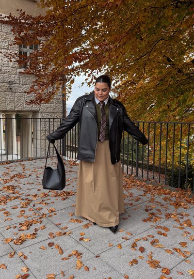 Chaqueta de cuero negra estilo biker, suéter verde, corbata con estampado, falda larga color beige, bolso negro, sobre un fondo de hojas otoñales y arquitectura de piedra.