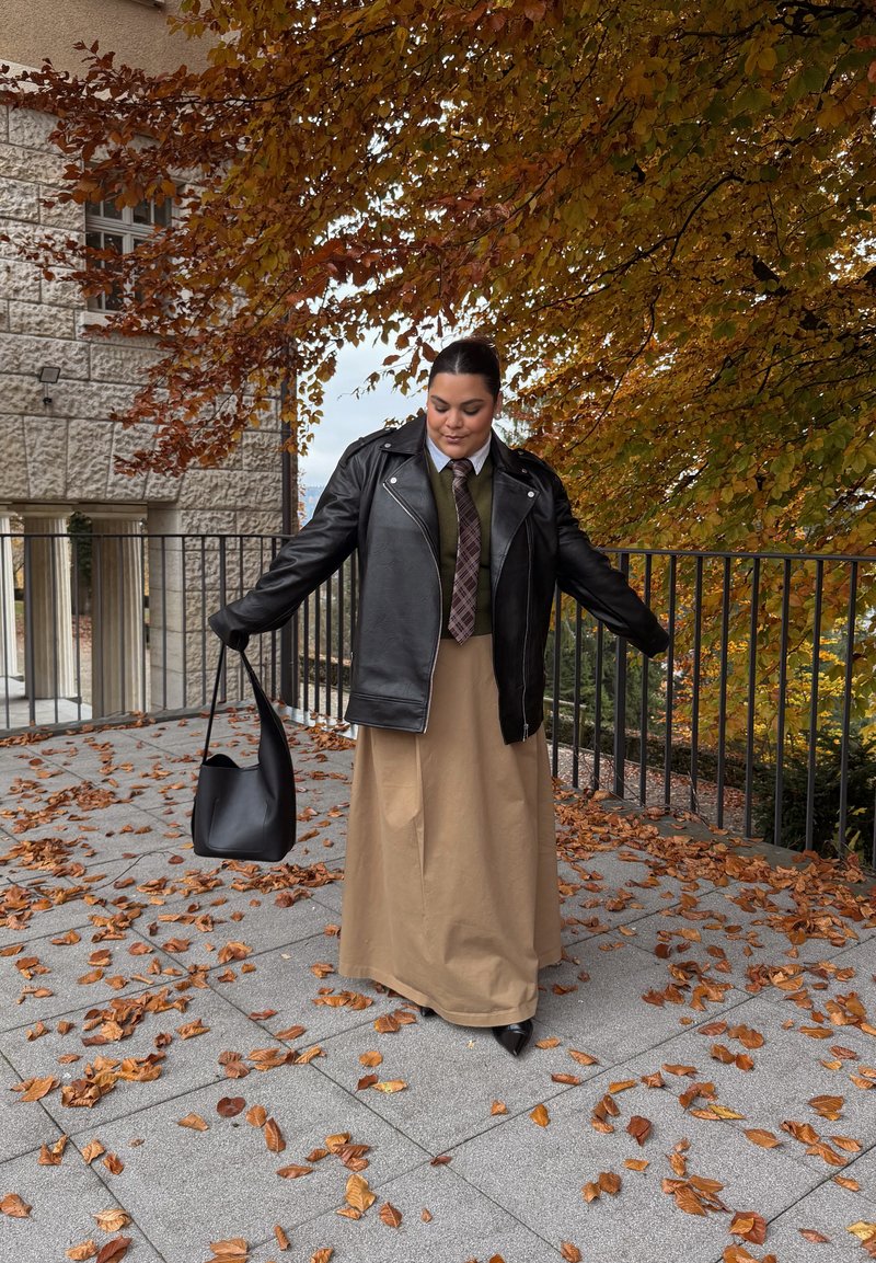 Schwarze Leder-Bikerjacke, grüner Pullover, gemusterte Krawatte, hellbrauner Maxirock, schwarze Handtasche, vor herbstlichen Blättern und steinerner Architektur.