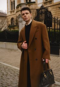 Manteau marron en laine à double boutonnage avec revers à pointe, associé à une chemise noire, tenant un sac à main en cuir noir avec des accents dorés.