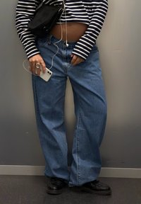 Kék, magasderekú, laza szabású farmernadrág fekete oldaltáskával és csíkos, hosszú ujjú felsővel párosítva. Tartozik hozzá egy iPhone és fülhallgató.