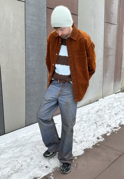 Hombre con un gorro beige, chaqueta de pana marrón, suéter a rayas, jeans azules y zapatillas, de pie sobre un pavimento nevado junto a una pared con paneles de metal.