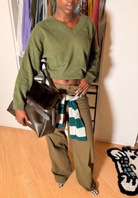 Άτομο που φοράει ελιά πράσινη κοντή μπλούζα και παντελόνια, κρατώντας μεγάλο σκούρο καφέ τσάντα, με ριγέ πουκάμισο δεμένο γύρω από τη μέση και παπούτσια με μοτίβο ζέβρας.