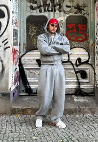 Szürke kapucnis pulcsi és hozzá illő melegítőnadrág, laza szabásban, piros sapkával és fehér sportcipővel viselve, graffiti-val borított fal előtt pózolva.