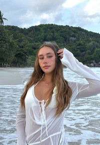 Top bianco trasparente a maniche lunghe con scollo profondo e dettagli con coulisse. Ambientazione tranquilla sulla spiaggia con onde e colline verdi sullo sfondo.