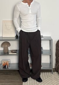 Chemise henley blanche à manches longues avec détails à boutons, associée à un pantalon large marron. La tenue comprend des chaussures noires et des accessoires minimalistes.