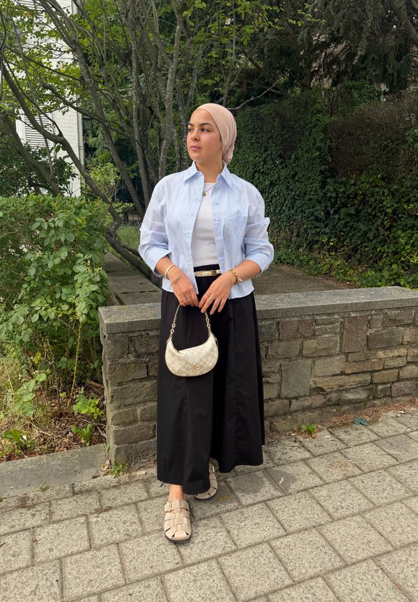 Femme portant un foulard beige, une chemise bleu clair, un top blanc et un pantalon noir à jambes larges, debout à l'extérieur, tenant un petit sac à main crème.