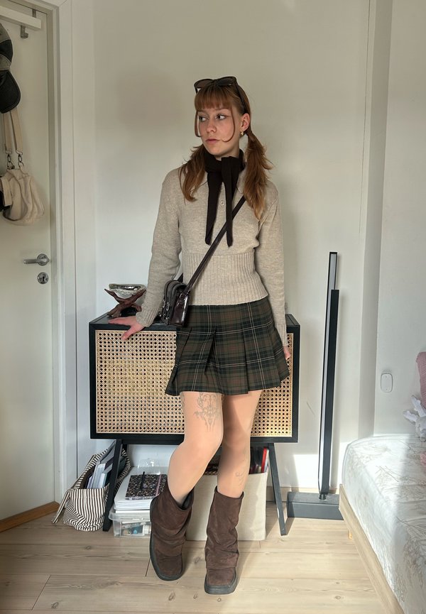 Une femme pose à l’intérieur, vêtue d’un pull beige, d’une jupe à carreaux, de bottes brunes et d’un sac en bandoulière, appuyée contre un meuble à façade en cannage.