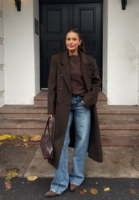 Donna con cappotto lungo marrone e maglione, jeans blu a gamba larga, che tiene una borsa marrone, in piedi vicino a gradini di pietra e una porta nera.