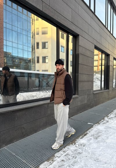 Hombre con chaleco marrón, gorro negro y pantalones gris claro caminando por una acera de rejilla metálica al lado de una calle nevada y ventanas de edificio reflectantes.