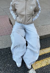 Pantaloni baggy grigi chiari con strisce bianche e una giacca beige con zip abbinata presentano un logo Adidas sul lato sinistro del petto. Tessuto morbido.