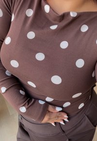 Braunes Langarmshirt mit weißen Punkten; strukturiertes Gewebe mit einem runden Ausschnitt. Modell hält sich an der Taille und trägt dunkle Hosen.