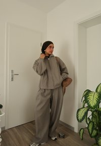 Veste zippée en polaire grise et pantalon large assorti, avec un sac à épaule marron et des baskets blanches ; en arrière-plan, une plante verte.