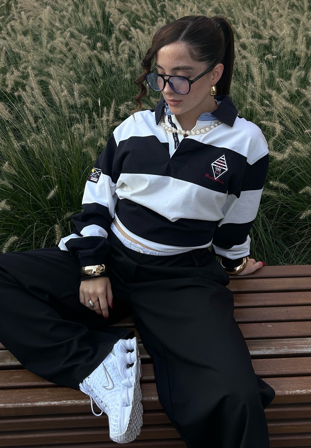 Černobíle pruhovaný ragbyový svetr s cropped designem, spárovaný s černými kalhotami a bílými teniskami. Obsahuje over-sized brýle a perlový náhrdelník.