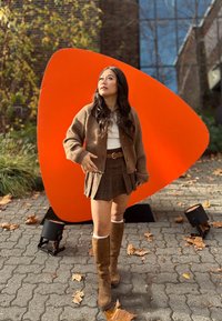 Cardigan marron en tricot, haut blanc, mini-jupe à carreaux, bottes hauteur genou couleur tan. En arrière-plan, une grande forme abstraite orange et du feuillage.