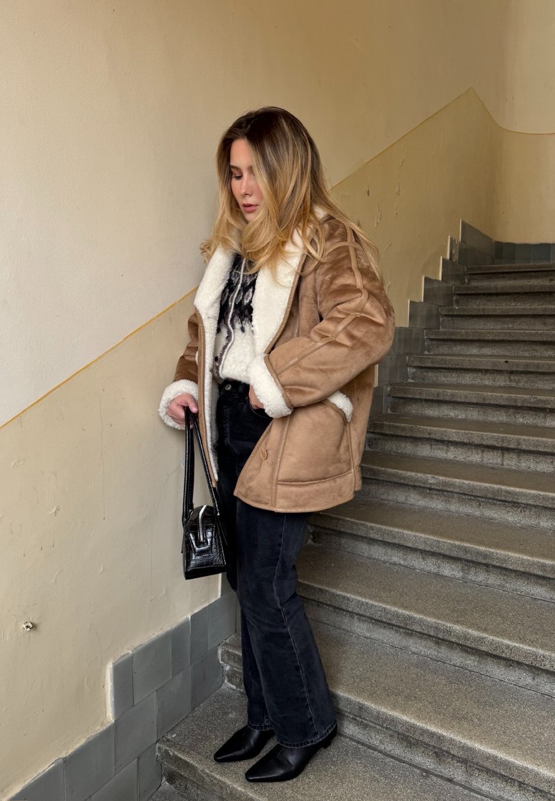 Femme en veste en peau lainée beige, pantalon noir et bottes, tenant un sac à main noir, debout sur des escaliers en pierre à l'intérieur.