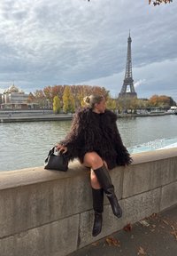 Femme en manteau de fourrure sombre et bottes assise sur un mur de pierre au bord de la rivière Seine avec la tour Eiffel et des arbres d'automne en arrière-plan.
