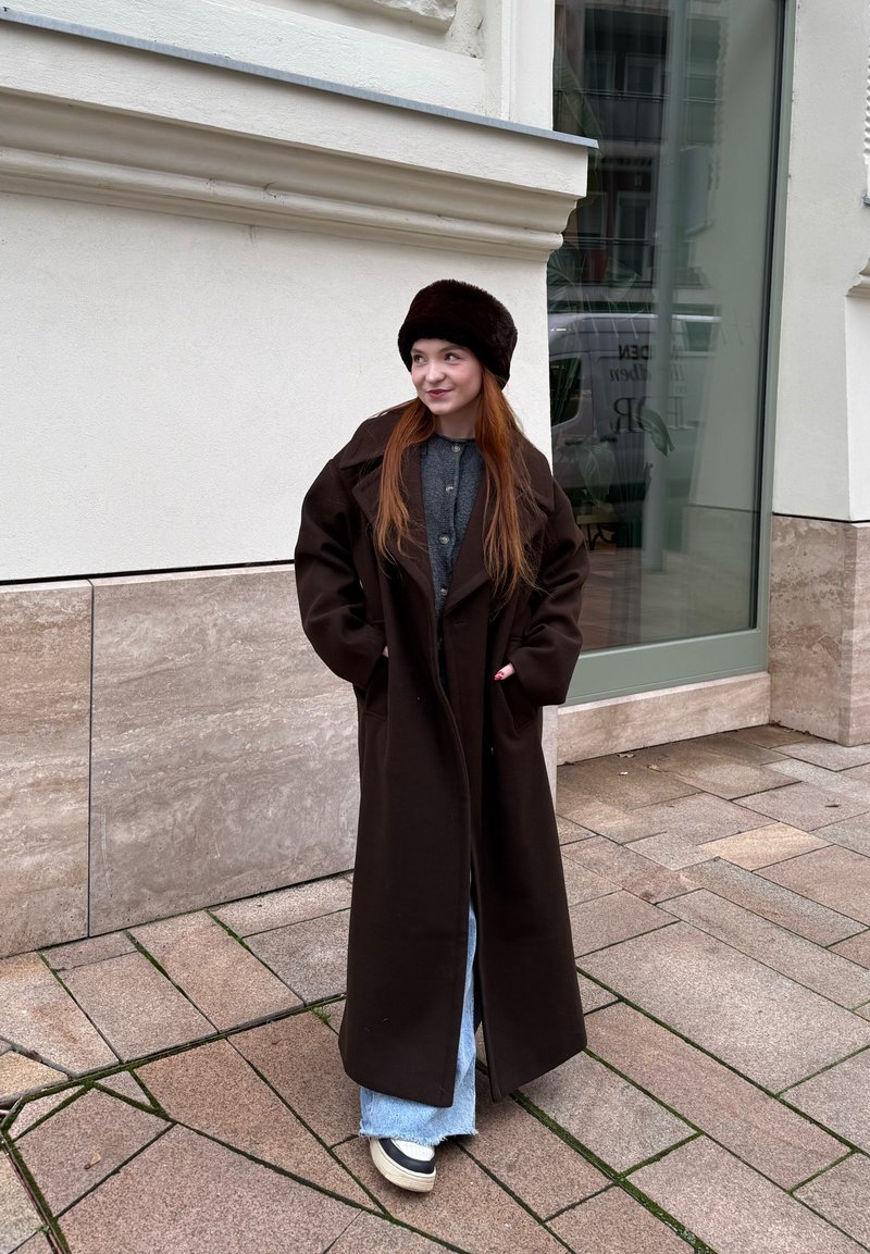 Femme aux longs cheveux roux portant un manteau marron foncé, un chapeau en fourrure, un jean et des baskets, debout sur un trottoir carrelé devant un bâtiment.