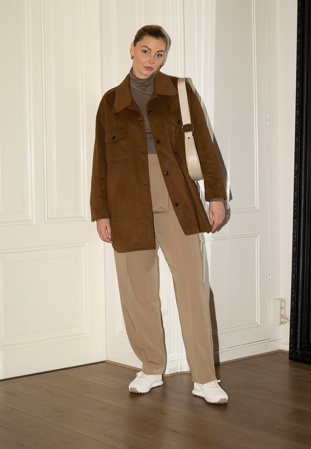 Casaco oversized em camurça castanha com dois bolsos no peito, suéter de gola alta bege, calças largas bege e ténis brancos. Chão de madeira clara, paredes brancas.