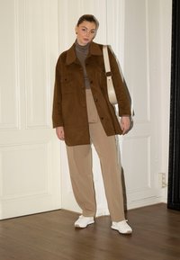 Casaco oversized em camurça castanha com dois bolsos no peito, suéter de gola alta bege, calças largas bege e ténis brancos. Chão de madeira clara, paredes brancas.